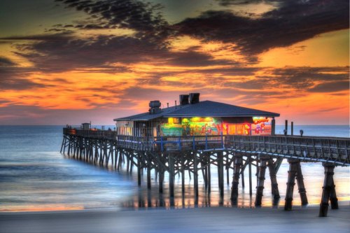 A colorful pier extends over calm waters at sunset, with vibrant clouds and a restaurant illuminated in the background.