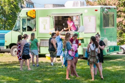 A green food truck serves customers in a grassy area, with people gathered around enjoying food and drinks.