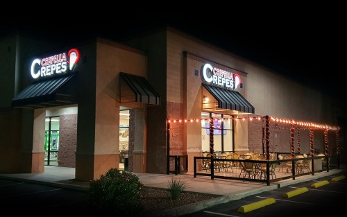 A nighttime view of a crepe restaurant with illuminated signage and outdoor seating decorated with string lights.