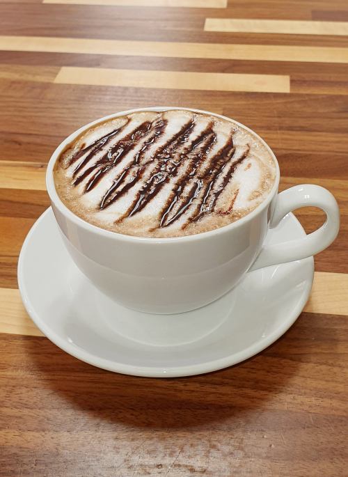 A white cup of coffee with chocolate drizzle on top, sitting on a saucer against a wooden table.