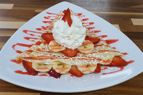 A festive crepe dessert shaped like a Christmas tree, topped with whipped cream, strawberries, and banana slices.