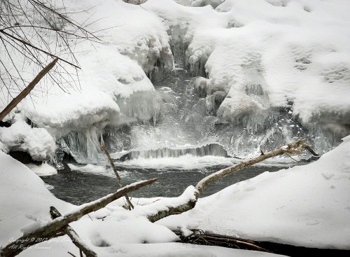 A frozen stream with ice formations and snow-covered banks, surrounded by bare branches.