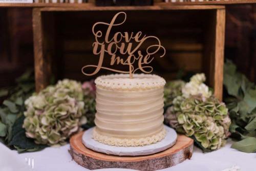 A beautifully decorated cake with a "Love You More" topper, surrounded by green hydrangeas on a rustic table.