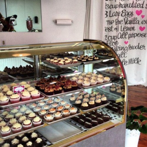 A display case filled with various cupcakes, with a recipe wall in the background featuring ingredients and measurements.