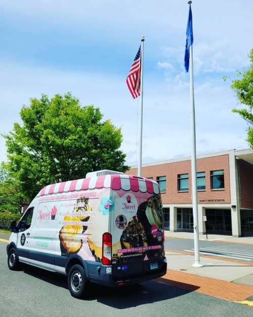 A colorful ice cream truck parked near a school, with flags flying in the background and trees lining the area.