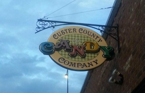 Sign for Custer County Candy Company, hanging from a brick building against a cloudy sky.