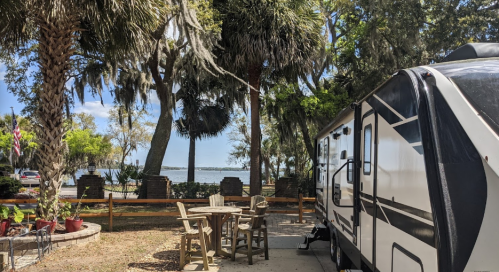 A scenic view of a campsite with a trailer, surrounded by palm trees and a lake in the background.