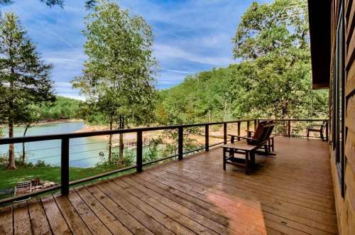 A wooden deck with two chairs overlooks a serene lake surrounded by lush greenery and trees under a blue sky.