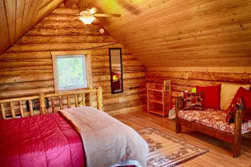 Cozy cabin interior with wooden walls, a bed with a red blanket, and a seating area by the window.