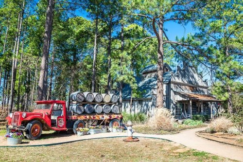 A vintage red truck with barrels parked in front of a rustic house surrounded by tall pine trees and colorful flowers.
