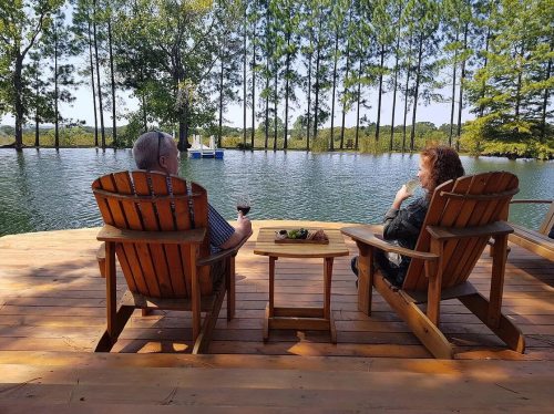 A couple relaxes in wooden chairs by a serene lake, enjoying drinks and the peaceful surroundings.