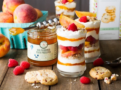 Two parfaits layered with yogurt, raspberries, and peach preserves, alongside fresh peaches and cookies on a wooden table.