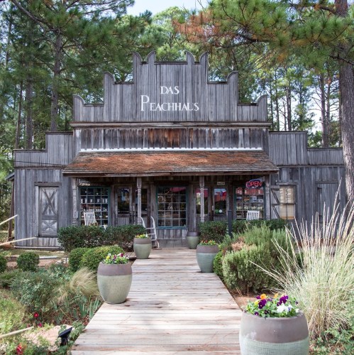 A rustic wooden building with "Das Peachhaus" sign, surrounded by greenery and flower pots, inviting entrance.