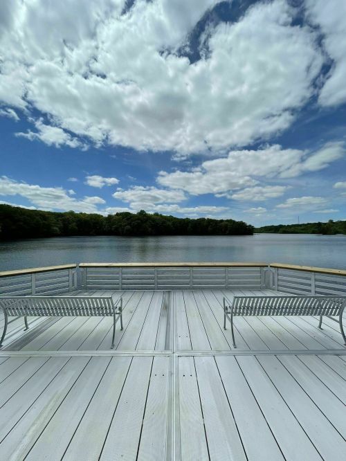 A serene view of a lake from a wooden deck, featuring two benches and a cloudy blue sky.