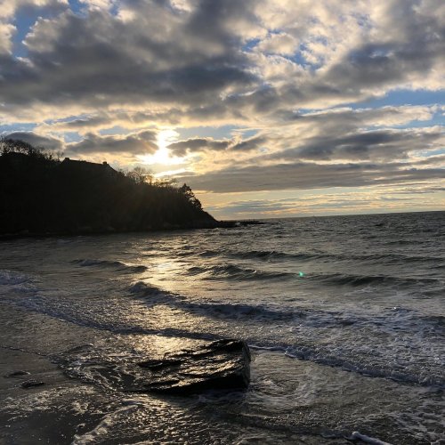 A serene beach scene at sunset, with clouds reflecting light over gentle waves and a rocky shore.