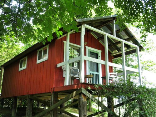 A small red cabin on stilts surrounded by greenery, featuring a porch and large windows.