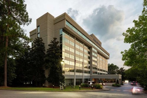 A modern hotel building with large windows, surrounded by trees, under a cloudy sky during twilight.