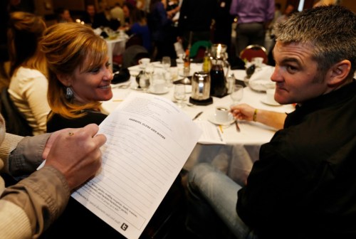 A man and woman sit at a dining table, while someone writes on a clipboard in the foreground.