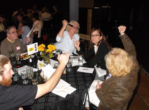 A group of people at a table, some raising their hands, with drinks and flowers in a lively setting.