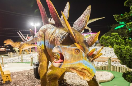 A colorful dinosaur statue with spikes, set in a mini-golf course at night, surrounded by rocks and greenery.