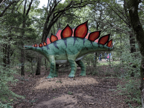 A large, colorful dinosaur statue resembling a stegosaurus stands among trees in a wooded area.