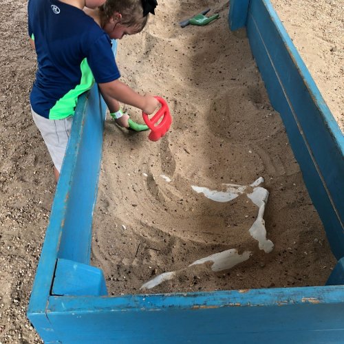 A child playing in a blue sandbox, using a red shovel to dig in the sand.