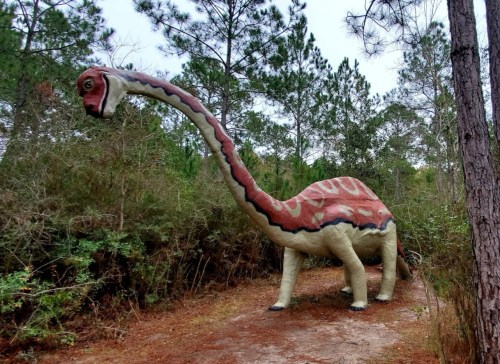 A large, colorful dinosaur statue stands among trees on a dirt path in a wooded area.