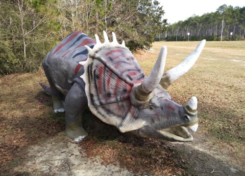A life-sized dinosaur sculpture of a Triceratops in a grassy area, surrounded by trees and a clear sky.
