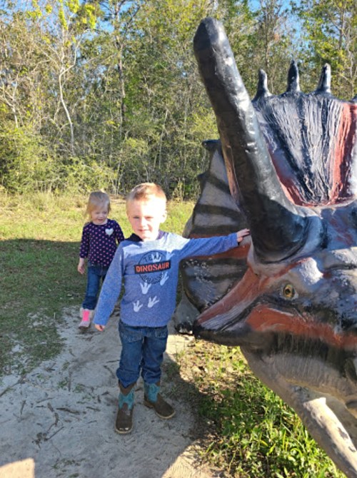 A boy in a dinosaur-themed shirt touches a dinosaur statue while a girl in the background walks nearby.