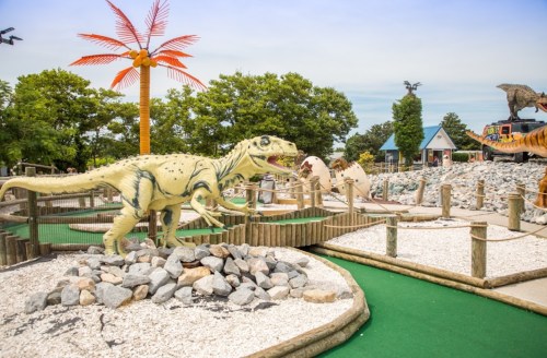 A mini-golf course featuring dinosaur sculptures, palm trees, and rocky landscaping under a clear blue sky.