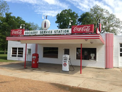 A vintage service station with Coca-Cola signage and retro gas pumps, surrounded by greenery.