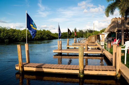 Wooden docks lined with flags along a calm river, surrounded by lush greenery and a blue sky with scattered clouds.
