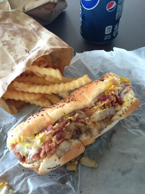 A hot dog topped with mustard, onions, and bacon, served with crinkle-cut fries and a can of Pepsi.