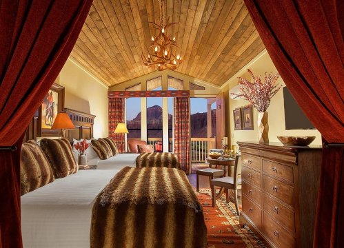 Cozy hotel room with two beds, wooden ceiling, warm decor, and mountain views through large windows.
