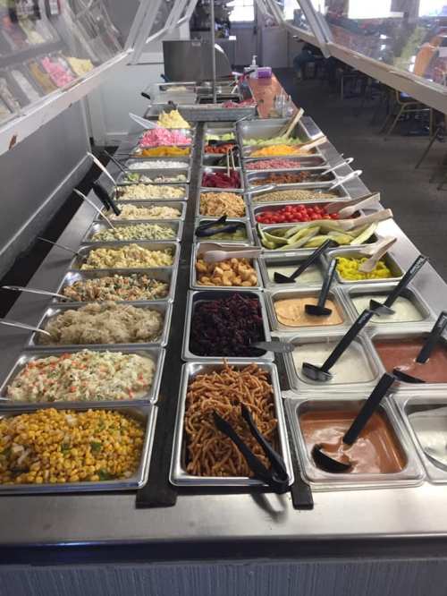 A buffet table filled with various salad toppings, including vegetables, dressings, and crunchy snacks in metal trays.