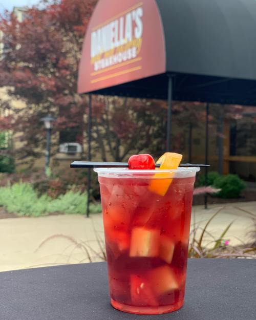 A refreshing fruit cocktail in a clear cup, garnished with a cherry and orange slice, outside a steakhouse.
