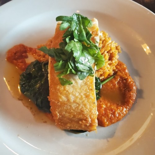 A plated dish featuring a piece of fish on a bed of greens, accompanied by a sauce and rice.