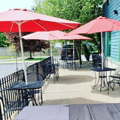 Outdoor seating area with black tables and chairs, shaded by red umbrellas, surrounded by greenery.