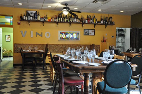 Cozy restaurant interior with a large wooden table set for dining, shelves lined with bottles, and a warm color scheme.