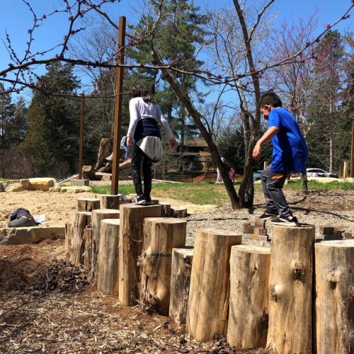 Two children balance on wooden logs in a playground, surrounded by trees and a sunny outdoor setting.