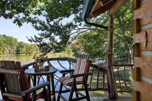 A cozy lakeside deck with wooden chairs, a table, and lush greenery surrounding a calm water view.