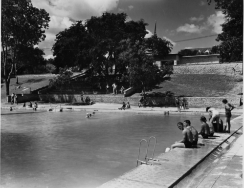 Black and white photo of a swimming pool with people lounging and swimming, surrounded by trees and grassy areas.