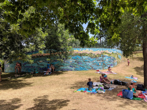 A colorful mural on a curved wall, with people relaxing on blankets in a grassy area under trees.