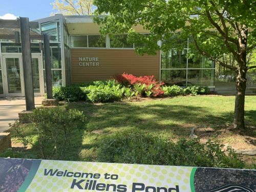 A nature center building surrounded by greenery and colorful plants, with a welcome sign for Killens Pond in the foreground.