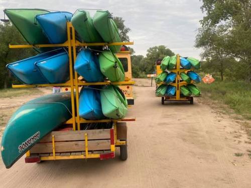 Two trailers loaded with stacked kayaks in green and blue, parked on a dirt road surrounded by greenery.