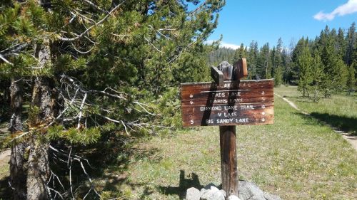 Wooden trail sign with directions to various lakes, surrounded by trees and a clear blue sky.