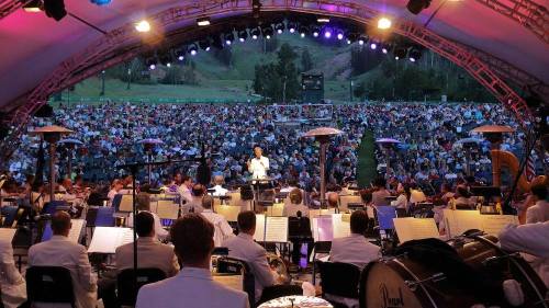 A conductor leads an orchestra in front of a large audience under a lit outdoor stage.