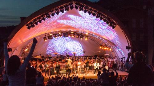 A lively outdoor concert with an orchestra and band performing under colorful lights, with an engaged audience.