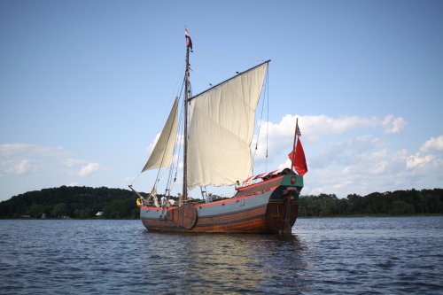 A wooden sailing ship with white sails and a red flag, anchored on calm water under a blue sky with clouds.