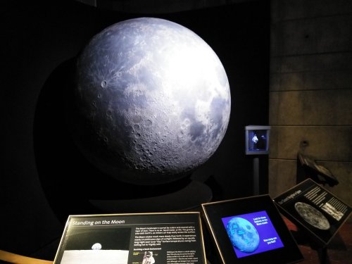 A large model of the Moon displayed in a museum, with informational plaques and a small screen showing lunar images.